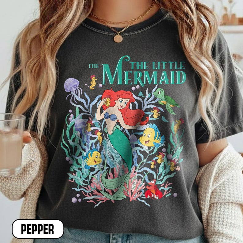 Puede incluir: Camiseta gris oscuro con un gr&aacute;fico colorido de Ariel de La Sirenita. El dise&ntilde;o incluye a Ariel, Flounder, Sebasti&aacute;n y otras criaturas marinas. El texto "THE LITTLE MERMAID" est&aacute; arqueado en la parte superior del dise&ntilde;o.