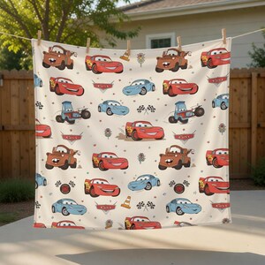 Puede incluir: Una tela color crema con un patr&oacute;n repetido de coches de dibujos animados en rojo, azul y marr&oacute;n. El dise&ntilde;o incluye banderas de carreras, conos de tr&aacute;fico y la palabra "Cars" en pancartas. Los coches tienen caras sonrientes.
