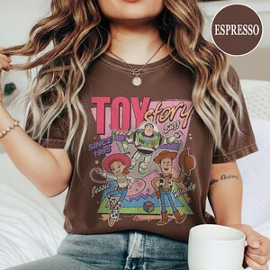 May include: Espresso-coloured t-shirt with a "Toy Story" graphic. The design features the film's title, characters Buzz Lightyear, Jessie, and Woody, and the text "Since 1995."
