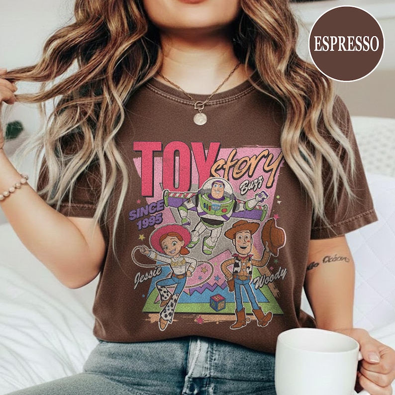 May include: Espresso-coloured t-shirt with a "Toy Story" graphic. The design features the film's title, characters Buzz Lightyear, Jessie, and Woody, and the text "Since 1995."
