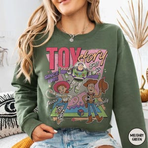 May include: Military green sweatshirt with a graphic of Toy Story characters Buzz Lightyear, Woody, and Jessie. The design includes the text "TOY STORY" and "SINCE 1995". The sweatshirt is a crew neck style.