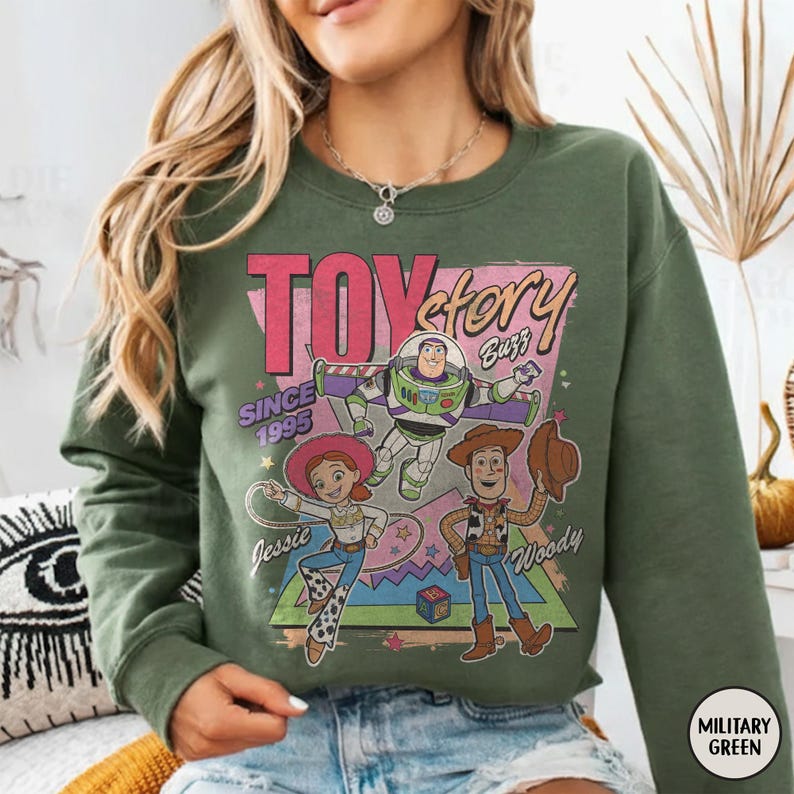 May include: Military green sweatshirt with a graphic of Toy Story characters Buzz Lightyear, Woody, and Jessie. The design includes the text "TOY STORY" and "SINCE 1995". The sweatshirt is a crew neck style.
