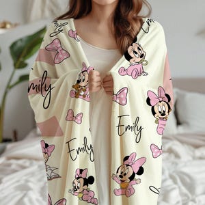 May include: Cream-colored blanket featuring a repeating pattern of Minnie Mouse in pink outfits, pink bows, and the name "Emily" in a script font. The blanket has pink accent stripes.