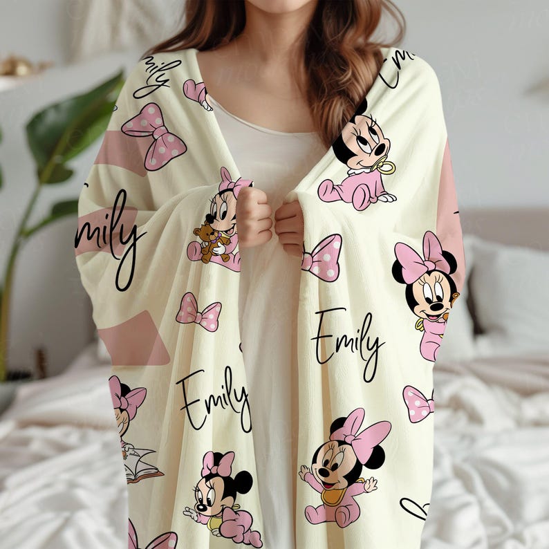 May include: Cream-colored blanket featuring a repeating pattern of Minnie Mouse in pink outfits, pink bows, and the name "Emily" in a script font. The blanket has pink accent stripes.