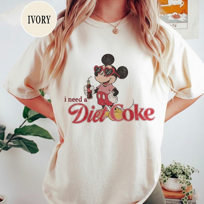 K&ouml;nnte beinhalten: Elfenbeinfarbenes T-Shirt mit einem Vintage-Mickey-Mouse-Motiv, das eine herzf&ouml;rmige Sonnenbrille tr&auml;gt und eine Diet Coke h&auml;lt. Der Text "i need a Diet Coke" ist in roter Schrift darunter gedruckt.