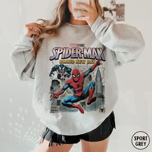 K&ouml;nnte beinhalten: Sportgraues Sweatshirt mit einem Marvel Studios Spider-Man-Motiv. Das Design zeigt Spider-Man, den Text "Brand New Day" und das Daily Bugle-Logo. Das Sweatshirt hat einen Rundhalsausschnitt und lange &Auml;rmel.