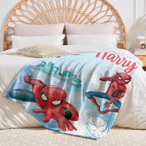 K&ouml;nnte beinhalten: Eine hellblaue Decke mit Illustrationen von Spider-Man in Actionposen. Die Decke hat ein Stadtbilddesign und den Namen "Harry" in roter Schrift. Die Decke liegt auf einem wei&szlig;en Bett.