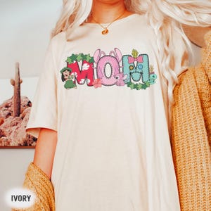 K&ouml;nnte beinhalten: Elfenbeinfarbenes T-Shirt mit dem Wort "MOM" in einem farbenfrohen Cartoon-Design. Die Buchstaben sind mit floralen und Charakterelementen verziert, darunter eine Hula-T&auml;nzerin, Hasenohren und eine Stitch-Figur. Das T-Shirt hat kurze &Auml;rmel.