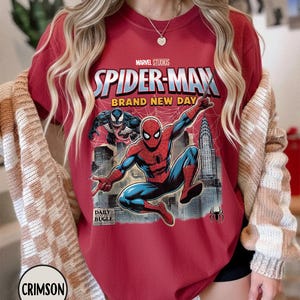 K&ouml;nnte beinhalten: Karmesinrotes T-Shirt mit einem Spider-Man-Motiv von Marvel Studios. Das Design zeigt Spider-Man in Aktion, den Text "SPIDER-MAN BRAND NEW DAY" und das Daily Bugle-Logo. Das T-Shirt hat einen Rundhalsausschnitt und kurze &Auml;rmel.
