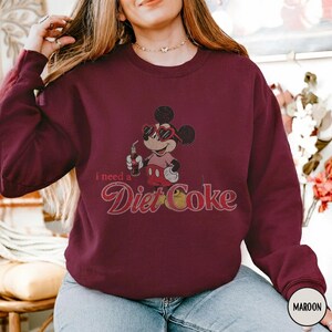May include: Maroon sweatshirt featuring a graphic of Mickey Mouse wearing sunglasses and holding a Diet Coke. The text "i need a Diet Coke" is printed below the graphic in a vintage style font. The sweatshirt has a crew neck.
