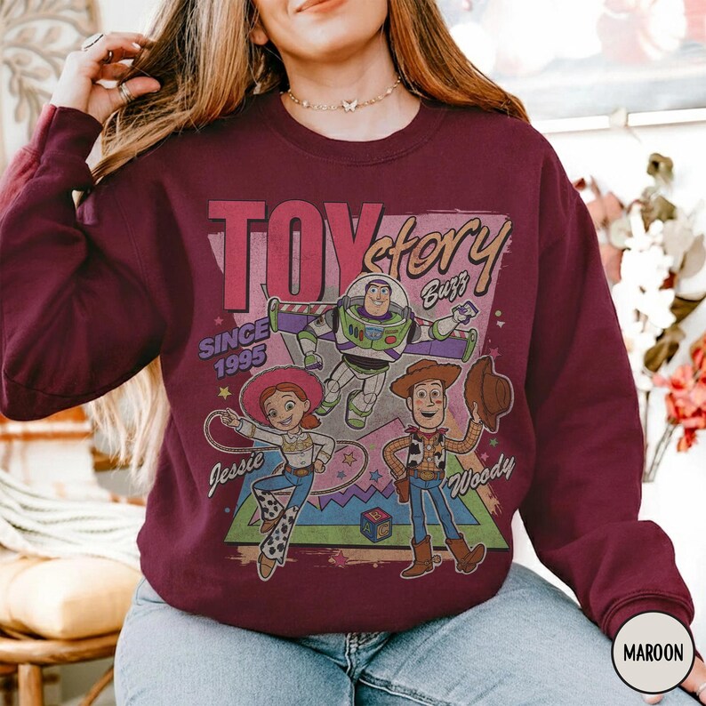 May include: Maroon sweatshirt with a "Toy Story" graphic. The design features Buzz Lightyear, Jessie, and Woody, with the text "Toy Story" and "Since 1995." The sweatshirt is a deep red colour, with a vintage-style design.