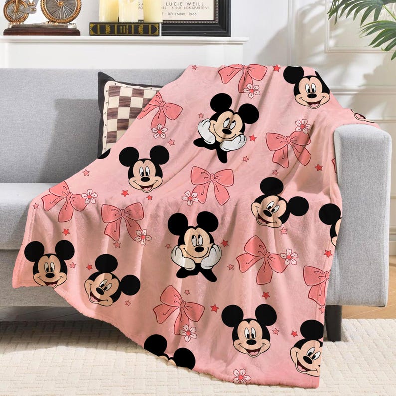 Op de afbeelding: Roze plaid met een herhalend patroon van Mickey Mouse, roze strikken, bloemen en rode sterren. De deken is over een grijze bank gedrapeerd en toont de zachte, pluche textuur en het levendige ontwerp.