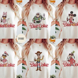 K&ouml;nnte beinhalten: Elfenbeinfarbene T-Shirts mit Cartoon-Charakteren und dem Text "I need a Diet Coke." Designs umfassen Daisy Duck, Buzz Lightyear, Mickey Mouse, Donald Duck, Woody und Goofy. Der Text "Diet Coke" ist in Schreibschrift.