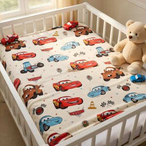 Puede incluir: Una s&aacute;bana de cuna color crema con un patr&oacute;n repetido de coches de dibujos animados en rojo, azul y marr&oacute;n. La s&aacute;bana est&aacute; en una cuna blanca, con coches de juguete y un oso de peluche cerca. El logotipo de Cars es visible.