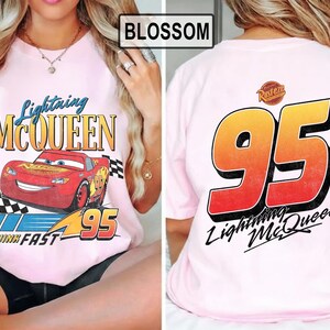 Pu&ograve; includere: T-shirt rosa chiaro con Lightning McQueen del film Cars. La parte anteriore presenta il personaggio e il testo, mentre il retro mostra un grande "95" e il testo "Lightning McQueen". La maglietta ha maniche corte.
