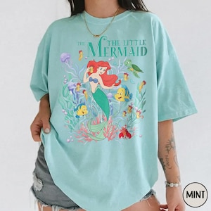 Puede incluir: Camiseta verde menta con un gr&aacute;fico colorido de "La Sirenita". El dise&ntilde;o incluye a Ariel, Flounder, Sebasti&aacute;n y otras criaturas marinas. El texto "The Little Mermaid" est&aacute; impreso encima del gr&aacute;fico.