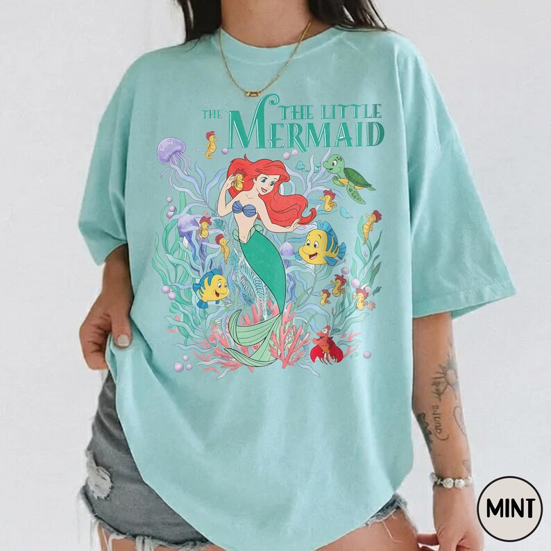 Puede incluir: Camiseta verde menta con un gr&aacute;fico colorido de "La Sirenita". El dise&ntilde;o incluye a Ariel, Flounder, Sebasti&aacute;n y otras criaturas marinas. El texto "The Little Mermaid" est&aacute; impreso encima del gr&aacute;fico.