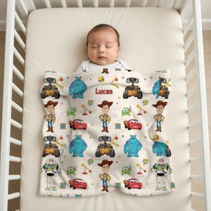 May include: A white baby blanket featuring cartoon characters from the movie Toy Story and Monsters, Inc. The blanket has images of Woody, Sully, Wall-E, Buzz Lightyear, and Lightning McQueen. The name "Lucas" is printed on the blanket.