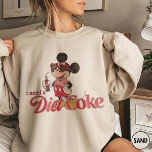 May include: Sand-colored sweatshirt featuring a vintage-style Mickey Mouse graphic. Mickey is wearing heart-shaped sunglasses and holding a Diet Coke. The text "i need a Diet Coke" is printed in red script.
