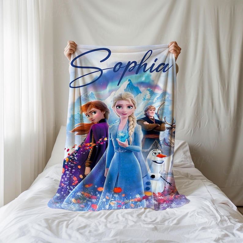 K&ouml;nnte beinhalten: Eine wei&szlig;e Fleece-Decke mit Figuren aus dem Film Frozen, darunter Elsa, Anna, Kristoff und Olaf. Die Decke hat den personalisierten Schriftzug "Sophia" in Blau. Der Hintergrund zeigt eine schneebedeckte Berglandschaft.