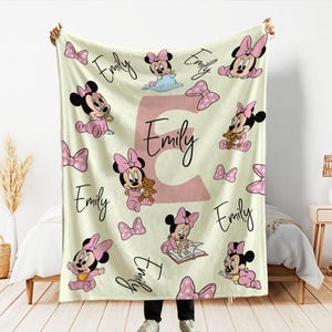 May include: A cream-colored baby blanket featuring Minnie Mouse illustrations in pink, along with pink bows and the name "Emily" in a script font. The letter "E" is prominently displayed in a matching pink hue.