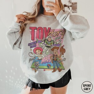 May include: Sport grey sweatshirt featuring a colorful graphic of Toy Story characters Buzz Lightyear, Woody, and Jessie. The design includes the text "Toy Story Buzz" and "Since 1995". The sweatshirt has a crew neck and long sleeves.