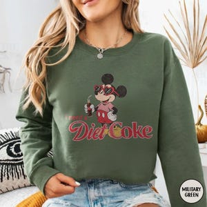 May include: Olive green sweatshirt featuring a vintage-style Mickey Mouse graphic wearing sunglasses and holding a Diet Coke. The text "I need a Diet Coke" is printed below the character in a retro font. The sweatshirt has a crew neck and long sleeves.