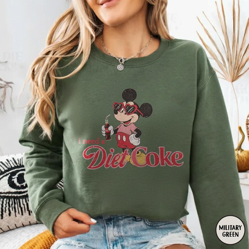 May include: Olive green sweatshirt featuring a vintage-style Mickey Mouse graphic wearing sunglasses and holding a Diet Coke. The text "I need a Diet Coke" is printed below the character in a retro font. The sweatshirt has a crew neck and long sleeves.