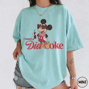 May include: A mint-colored t-shirt featuring a graphic of Mickey Mouse wearing sunglasses and holding a Diet Coke bottle. The text "i need a Diet Coke" is printed below the cartoon character in a retro font. The shirt has short sleeves and a relaxed fit.