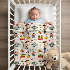 May include: A baby sleeps in a white crib, covered by a blanket featuring Toy Story characters. The blanket has a cream background with images of Woody, Buzz Lightyear, Mr. Potato Head, and the Toy Story logo. A teddy bear rests beside the crib.
