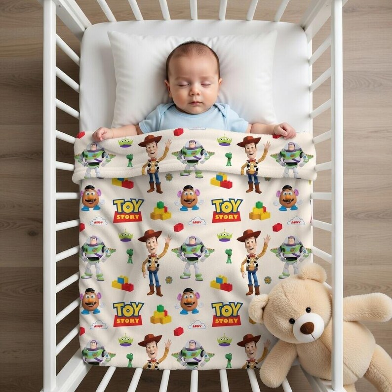 May include: A baby sleeps in a white crib, covered by a blanket featuring Toy Story characters. The blanket has a cream background with images of Woody, Buzz Lightyear, Mr. Potato Head, and the Toy Story logo. A teddy bear rests beside the crib.