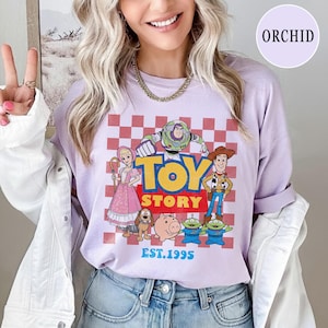 以下が含まれることがあります： バズ・ライトイヤーやウッディなどのキャラクターが描かれた、赤と白のチェック柄の背景に、TOY STORYのグラフィックがプリントされたライラック色の長袖シャツ。テキスト「TOY STORY」と「EST.1995」も表示されています。
