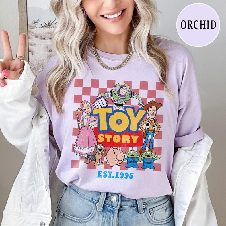 以下が含まれることがあります： バズ・ライトイヤーやウッディなどのキャラクターが描かれた、赤と白のチェック柄の背景に、TOY STORYのグラフィックがプリントされたライラック色の長袖シャツ。テキスト「TOY STORY」と「EST.1995」も表示されています。