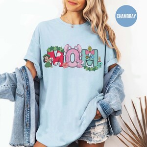 K&ouml;nnte beinhalten: Hellblaues T-Shirt mit dem Wort "MOM" in einem farbenfrohen Grafikdesign. Das "M" ist rot mit gr&uuml;nen Bl&auml;ttern, das "O" ist rosa mit Hasenohren und das "M" ist blau mit einer Schleife. Das Shirt hat einen l&auml;ssigen Stil.