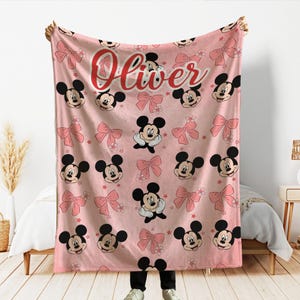 Op de afbeelding: Roze deken met een patroon van Mickey Mouse-gezichten, roze strikken en kleine bloemen. De naam "Oliver" staat in rode letters bovenaan. De deken wordt door een persoon omhoog gehouden.