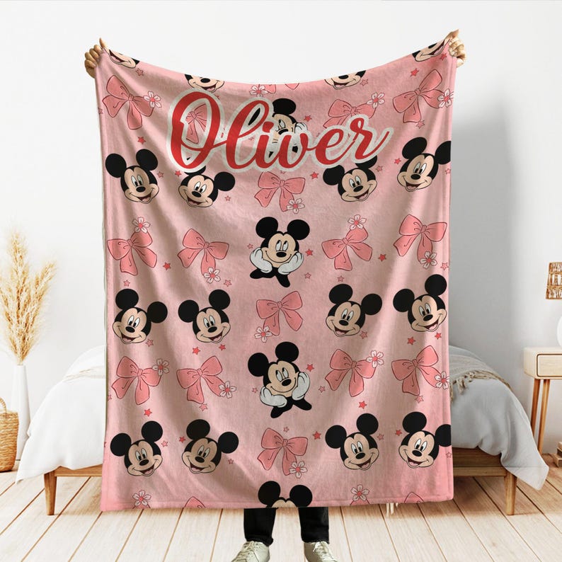 Op de afbeelding: Roze deken met een patroon van Mickey Mouse-gezichten, roze strikken en kleine bloemen. De naam "Oliver" staat in rode letters bovenaan. De deken wordt door een persoon omhoog gehouden.