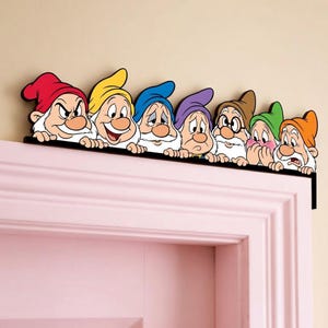 May include: A decorative door topper featuring seven cartoon dwarves with white beards and colourful hats in red, yellow, blue, purple, brown, green, and orange. Each character has a distinct facial expression and rests their hands on a black ledge.
