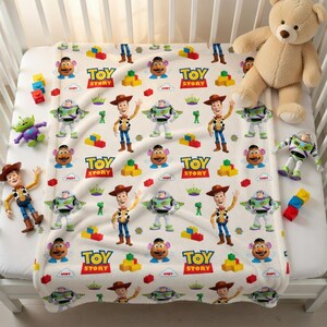 May include: A white baby blanket featuring a repeating pattern of Toy Story characters, including Woody, Buzz Lightyear, and Mr. Potato Head. The blanket is surrounded by Toy Story figurines and a teddy bear. The blanket is on a white crib.