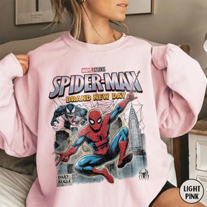 K&ouml;nnte beinhalten: Hellrosa Spider-Man-Sweatshirt mit einer Grafik von Spider-Man, Venom und dem Text "SPIDER-MAN BRAND NEW DAY". Das Daily Bugle-Logo ist ebenfalls sichtbar. Das Sweatshirt ist ein Rundhals-Stil.