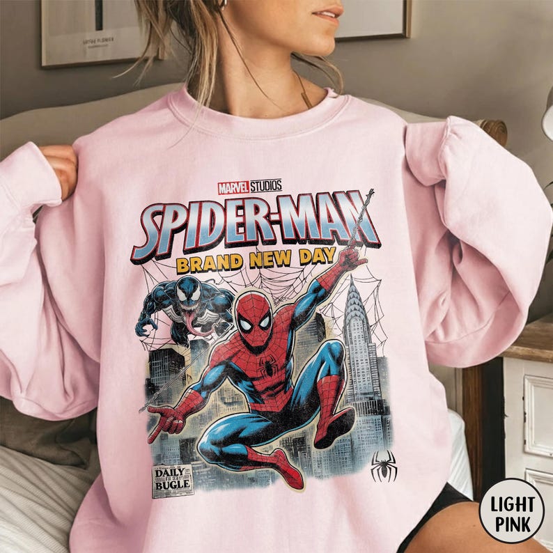 K&ouml;nnte beinhalten: Hellrosa Spider-Man-Sweatshirt mit einer Grafik von Spider-Man, Venom und dem Text "SPIDER-MAN BRAND NEW DAY". Das Daily Bugle-Logo ist ebenfalls sichtbar. Das Sweatshirt ist ein Rundhals-Stil.