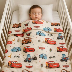 Puede incluir: Un beb&eacute; durmiendo en una cuna blanca, cubierto con una manta color crema. La manta presenta un patr&oacute;n repetido de coches de dibujos animados en rojo, azul y marr&oacute;n, junto con banderas de carreras y sem&aacute;foros. El beb&eacute; descansa sobre una almohada blanca.
