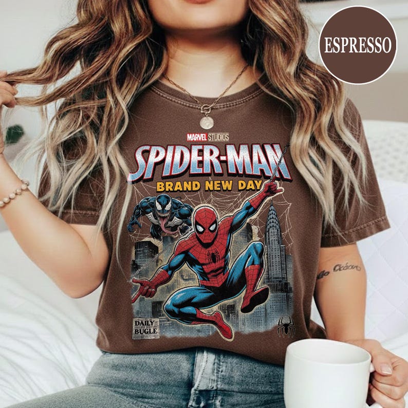 K&ouml;nnte beinhalten: Braunes T-Shirt mit einem Spider-Man-Motiv von Marvel Studios. Das Design zeigt Spider-Man in Aktion, den Text &bdquo;BRAND NEW DAY&ldquo; und das Daily Bugle-Logo. Das T-Shirt hat einen Rundhalsausschnitt und ist dunkelbraun.