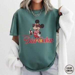 May include: Light green t-shirt featuring a graphic of Mickey Mouse wearing sunglasses and holding a Diet Coke bottle. The text "I need a Diet Coke" is printed below the character in a retro font. The shirt is a solid color.