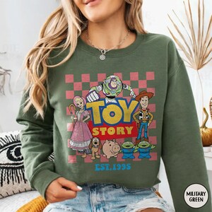 以下が含まれることがあります： バズ・ライトイヤー、ウッディなどのキャラクターが描かれた、ミリタリーグリーンのスウェットシャツ。レトロなデザインで「TOY STORY」と「EST. 1995」の文字が入っています。クルーネックのスウェットシャツです。