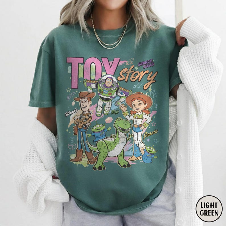 May include: Light green t-shirt featuring the words "TOY STORY" in pink and white, with the phrase "SINCE 1995." The shirt displays cartoon images of Woody, Buzz Lightyear, Jessie, and other characters from the movie. The shirt is a light green color.