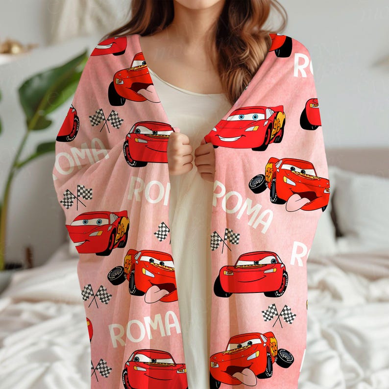 Puede incluir: Una manta rosa claro con un patr&oacute;n repetido del coche de carreras rojo Rayo McQueen de la pel&iacute;cula Cars, banderas a cuadros y la palabra "ROMA" en blanco. La manta parece suave y c&oacute;moda.