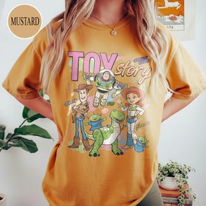 May include: Mustard-colored t-shirt featuring a graphic of characters from the animated film "Toy Story." The design includes Woody, Buzz Lightyear, Jessie, and other characters, with the text "TOY STORY SINCE 1995."
