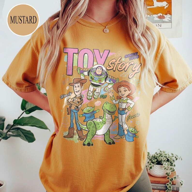 May include: Mustard-colored t-shirt featuring a graphic of characters from the animated film "Toy Story." The design includes Woody, Buzz Lightyear, Jessie, and other characters, with the text "TOY STORY SINCE 1995."