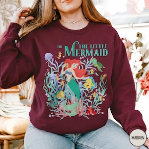 Puede incluir: Sudadera granate con un gr&aacute;fico colorido de Ariel de La Sirenita. El dise&ntilde;o incluye a Ariel, Flounder, Sebasti&aacute;n y otras criaturas marinas, con el texto "The Little Mermaid" en verde azulado.