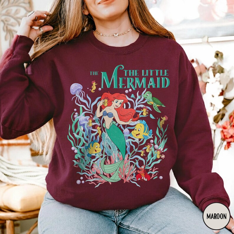 Puede incluir: Sudadera granate con un gr&aacute;fico colorido de Ariel de La Sirenita. El dise&ntilde;o incluye a Ariel, Flounder, Sebasti&aacute;n y otras criaturas marinas, con el texto "The Little Mermaid" en verde azulado.